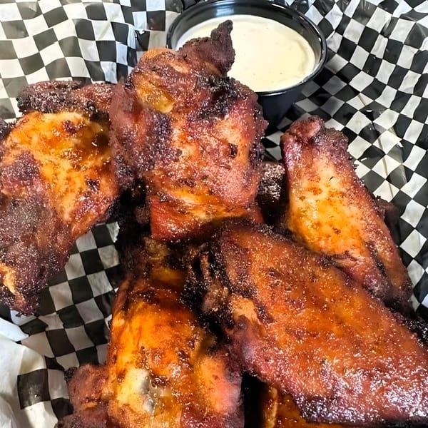 Close-up of several crispy, glazed chicken wings with a side of ranch dressing on checkered paper.