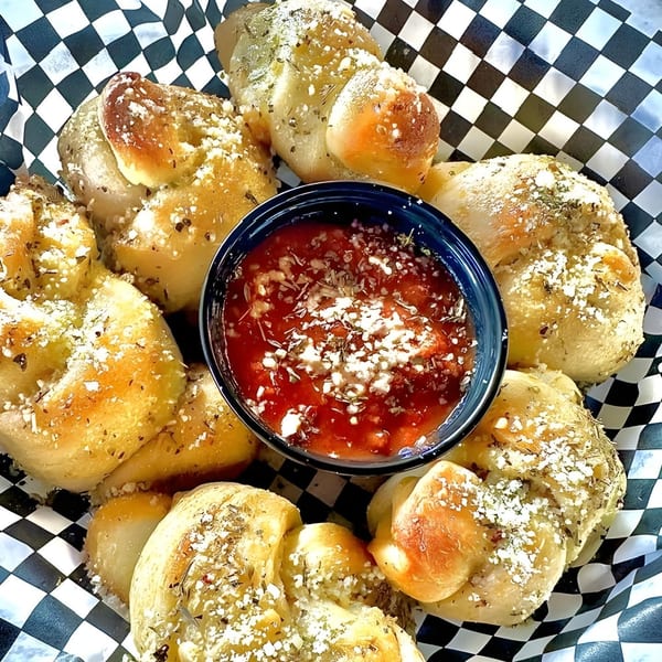 Garlic knots with parmesan cheese and herbs, served with a side of marinara sauce in a checkered basket.