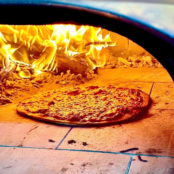 A Baby Hughy's pizza baking inside a wood-fired oven with bright flames in the background.