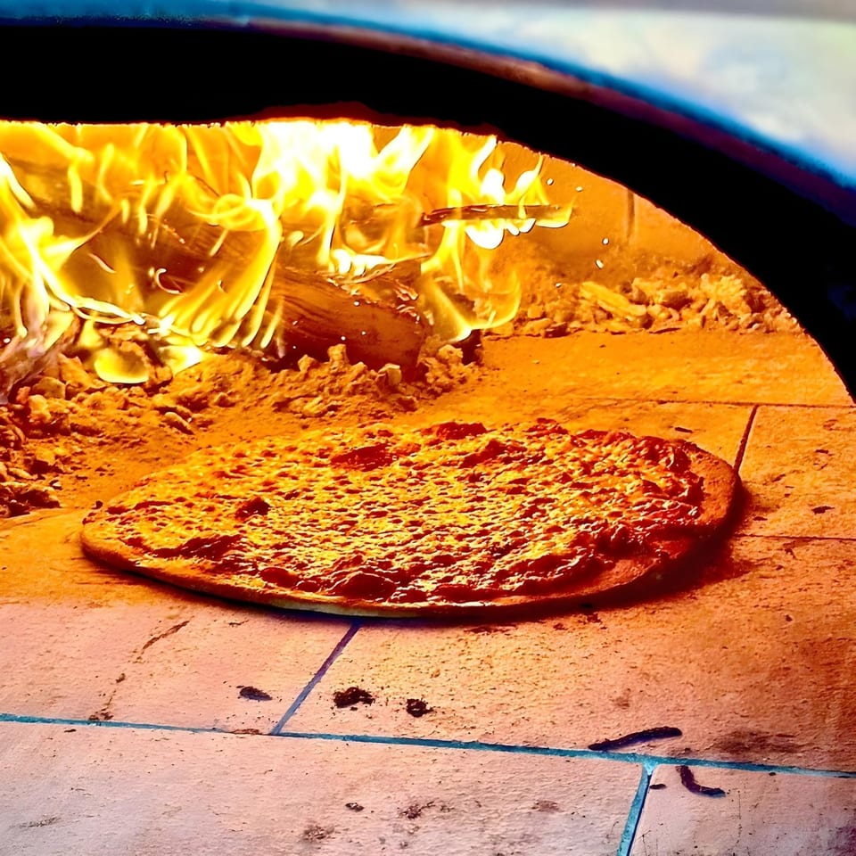 A Baby Hughy's pizza baking inside a wood-fired oven with bright flames in the background.