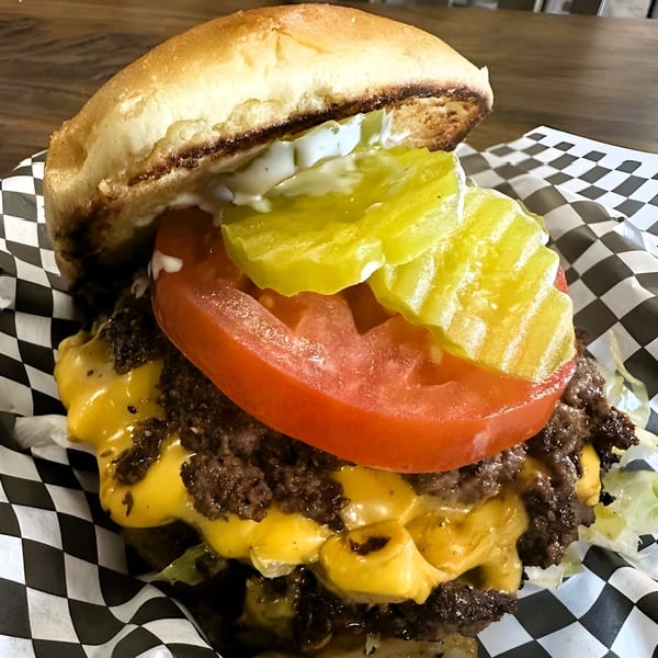 A close-up of a double cheeseburger with lettuce, tomato, pickles, and sauce on checkered paper.