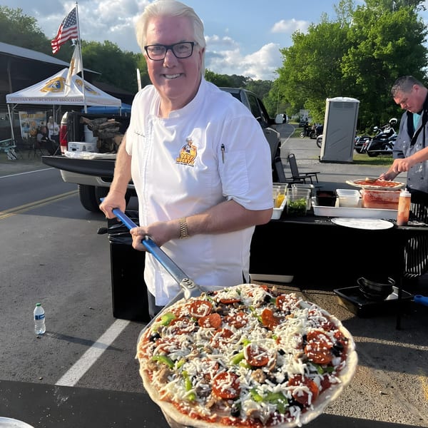A chef from Baby Hughy's Restaurant holds a large, uncooked pizza with various toppings on a peel.