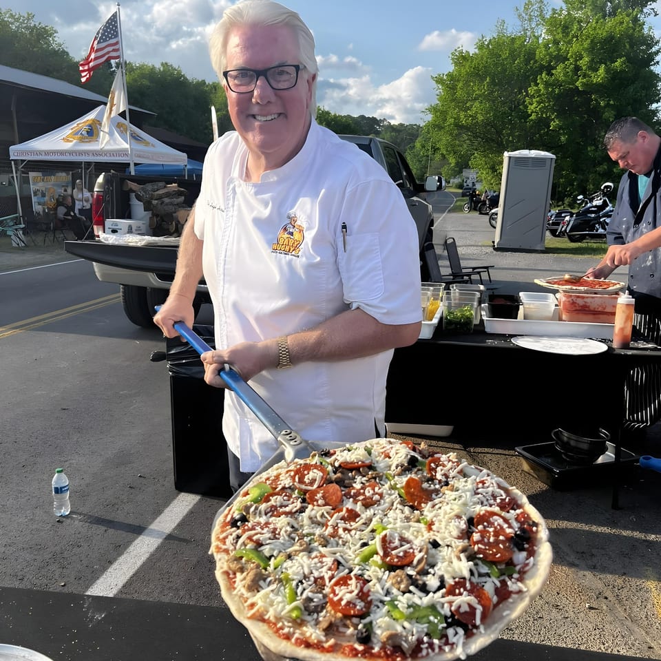 A chef from Baby Hughy's Restaurant holds a large, uncooked pizza with various toppings outdoors.