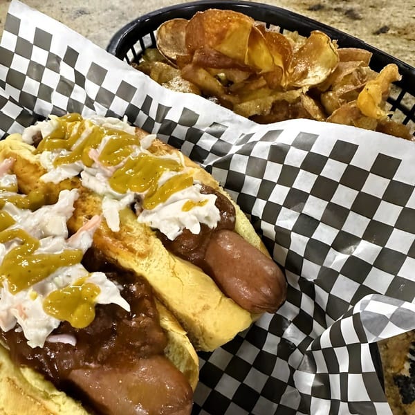 Two hot dogs with chili, coleslaw, and mustard, served with a side of potato chips.